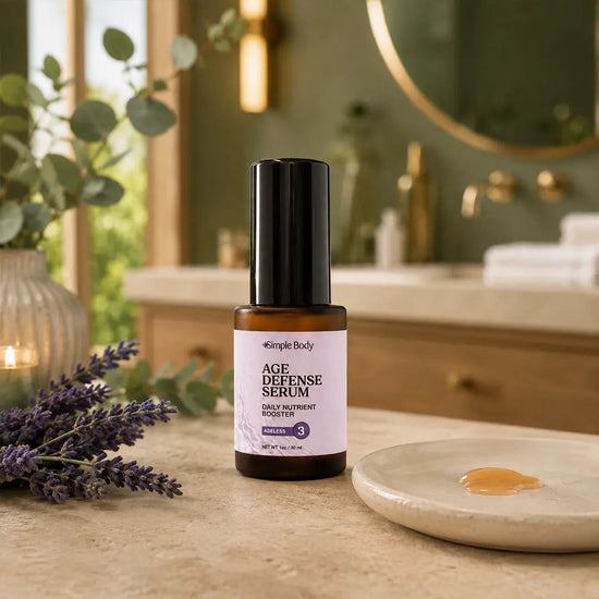 A bottle of Simple Body Age Defense Serum stands on a bathroom counter with a small dish of serum, lavender sprigs, and a blurred vanity in the background.
