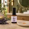 A bottle of Simple Body Age Defense Serum stands on a bathroom counter with a small dish of serum, lavender sprigs, and a blurred vanity in the background.