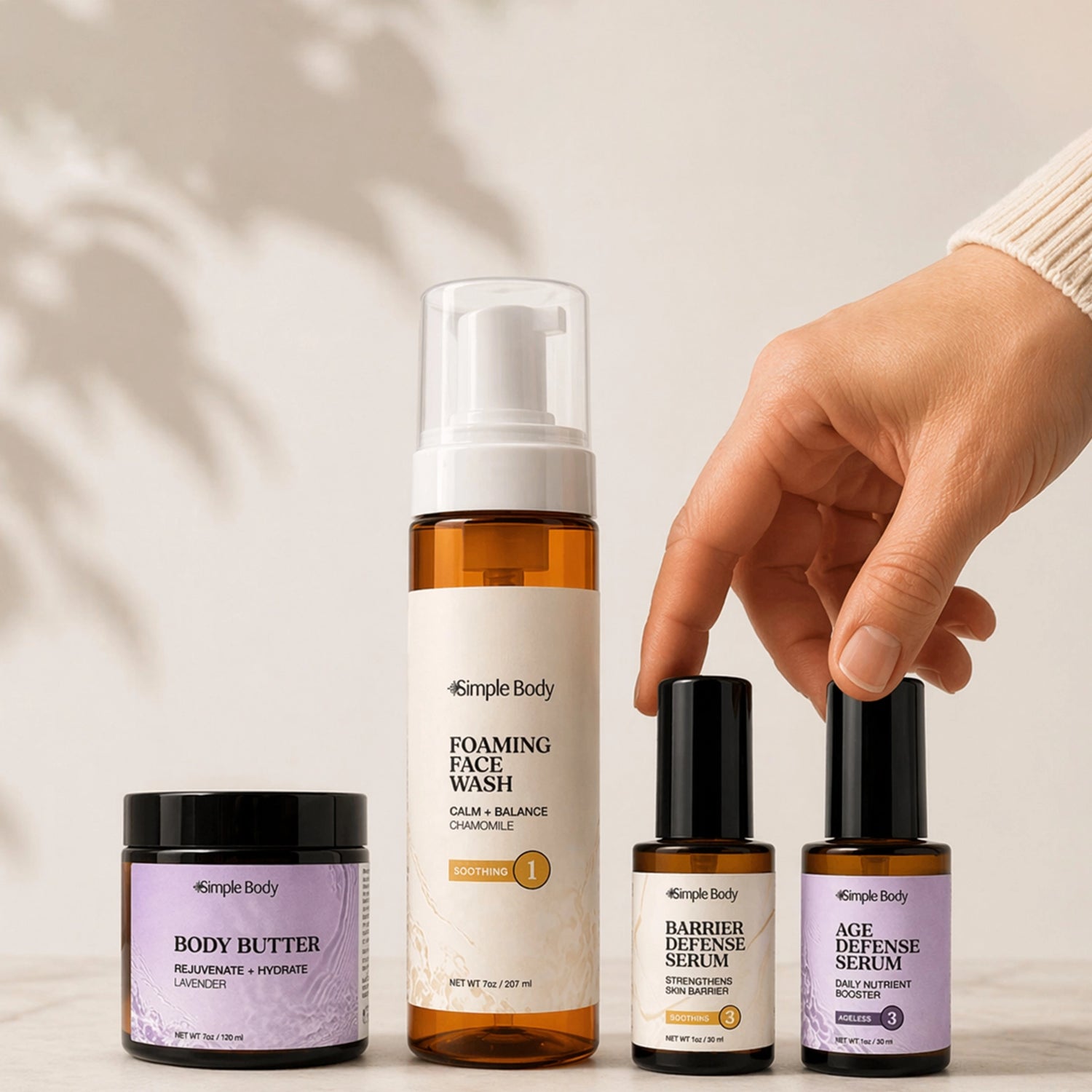 A woman's hand grabs for a Simple Body Barrier Defense Serum, Age Defense Serum, Foaming Face Wash in Chamomile and a Lavender Body Butter with a beige background and shadow from a plant.