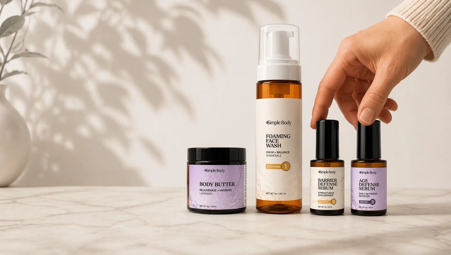 A woman's hand grabs for a Simple Body Barrier Defense Serum, Age Defense Serum, Foaming Face Wash in Chamomile and a Lavender Body Butter with a beige background and shadow from a plant.