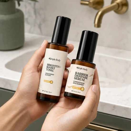 Two hands holding two amber glass skincare bottles one a Brighten + Boost Tonic and a Barrier Defense Serum with black caps from the brand Simple Body, set against a bathroom sink background with a gold faucet.