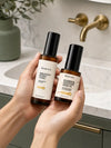 Two hands holding two amber glass skincare bottles one a Brighten + Boost Tonic and a Barrier Defense Serum with black caps from the brand Simple Body, set against a bathroom sink background with a gold faucet.