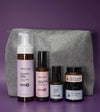A Starter Skincare Kit for mature skin with a foaming face wash, Rosewater Toner, Nightly Face Serum, Beauty Balm, Face Cream, and a gray cosmetic bag on a purple background.