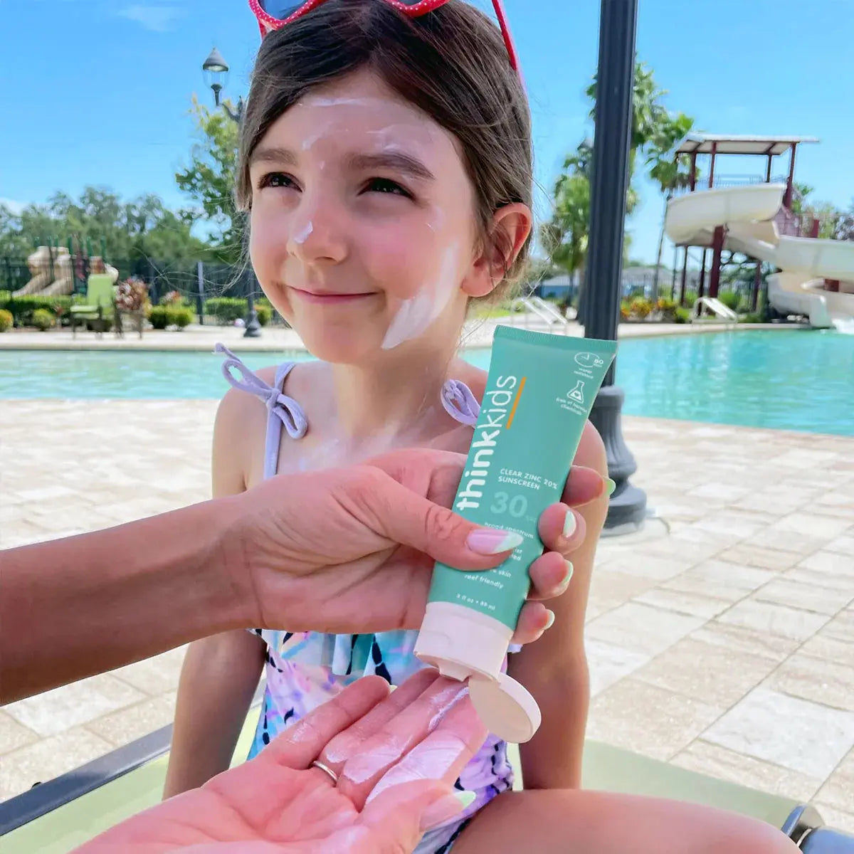 Child at a poolside with sunscreen being applied, holding a tube of thinkkids sunscreen.