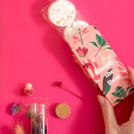 A photo of a hand dispensing a Lily and Rose Bath Steamer Two Pack from a box that is pink with beautiful floral illustrations that reads: Simply the Best along with some fresh dried botanicals on a hot pink background.