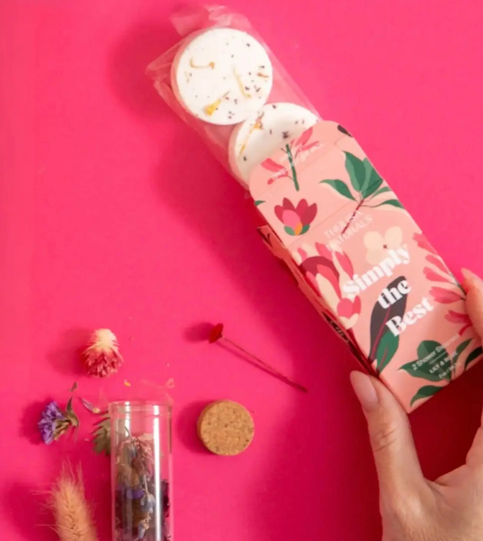 A photo of a hand dispensing a Lily and Rose Bath Steamer Two Pack from a box that is pink with beautiful floral illustrations that reads: Simply the Best along with some fresh dried botanicals on a hot pink background.