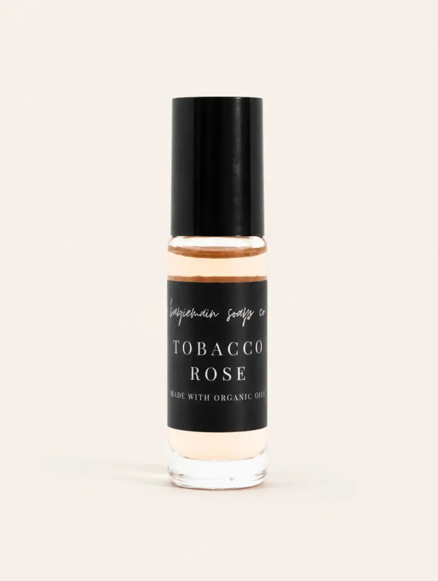 A small glass bottle of Robacco Rose Perfume Oil from Benjamin Soap Co with a black cap and black label, containing a warm amber liquid, centered on a light cream background.