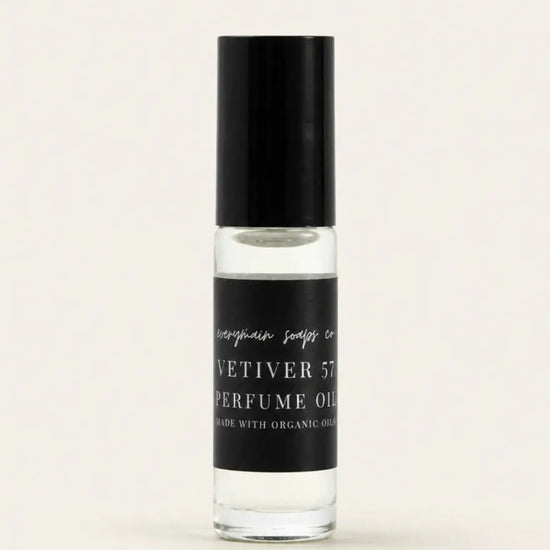 A small clear glass bottle of Vetiver 57 perfume oil with a black cap and a black label, centered on a warm off-white background.