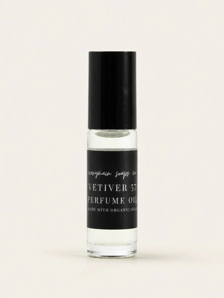 A small clear glass bottle of Vetiver 57 perfume oil with a black cap and a black label, centered on a warm off-white background.