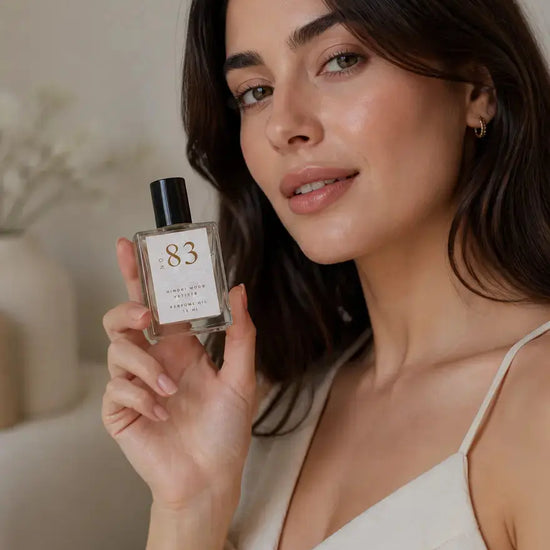 A woman holds a bottle of Luxe Perfume oil as the label reads "no. 83 Hinoki Wood Vetiver Perfume Oil on a beige background.