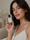 A woman holds a bottle of Luxe Perfume oil as the label reads "no. 83 Hinoki Wood Vetiver Perfume Oil on a beige background.