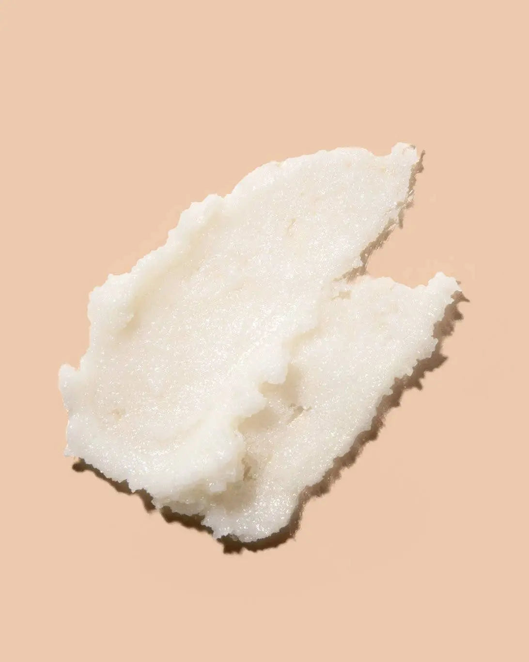 A dollop of white creamy body scrub photographed on a solid peach-beige background.