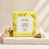 A box of LUMI Lemon Ginger herbal tea sits on a light stone surface with a lemon wedge, ginger root, and a pyramid tea bag beside it. The box is bright yellow with black botanical line art and a white front label.