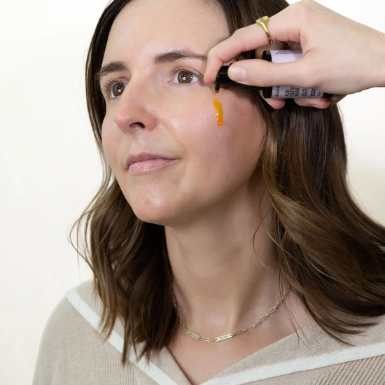 Woman applying Simple Body Age Defense Serum which is orange in color to her cheek white background