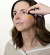 Woman applying Simple Body Age Defense Serum which is orange in color to her cheek white background