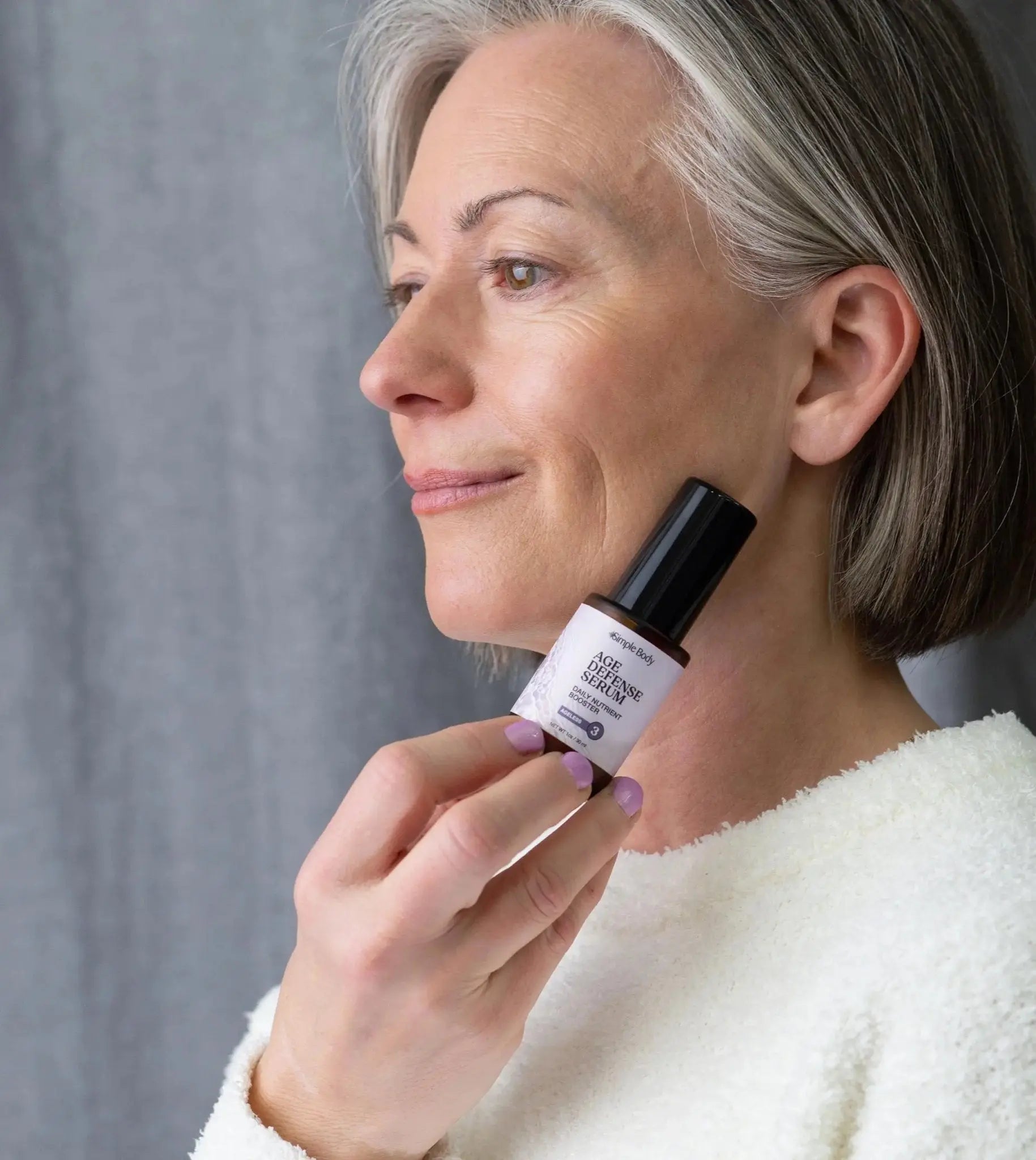 Michelle holding a bottle of Simple Body Age Defense Serum near her cheek with purple fingernails agains a gray backdrop.