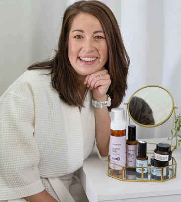Woman sitting with a Simple Body Age Supporting Skincare Kit including Foaming Face Wash, Age Defying Tonic, Wrinkle Defense Serum, Face Cream, and Beauty Balm in a little gold tray with a mirror on a white background.