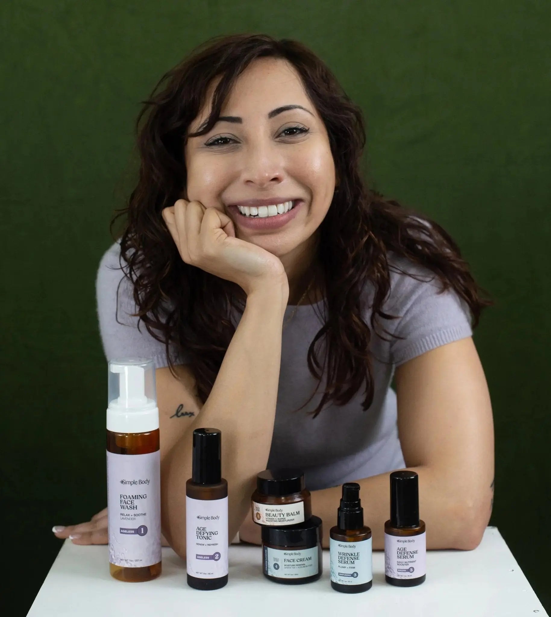 Saba sitting in front of a green backdrop smiling with Simple Body Foaming Face Wash, Age Defying Tonic, Beauty Balm, Face Cream, Wrinkle Defense, and Age Defense Serum in front of her on a white stand.