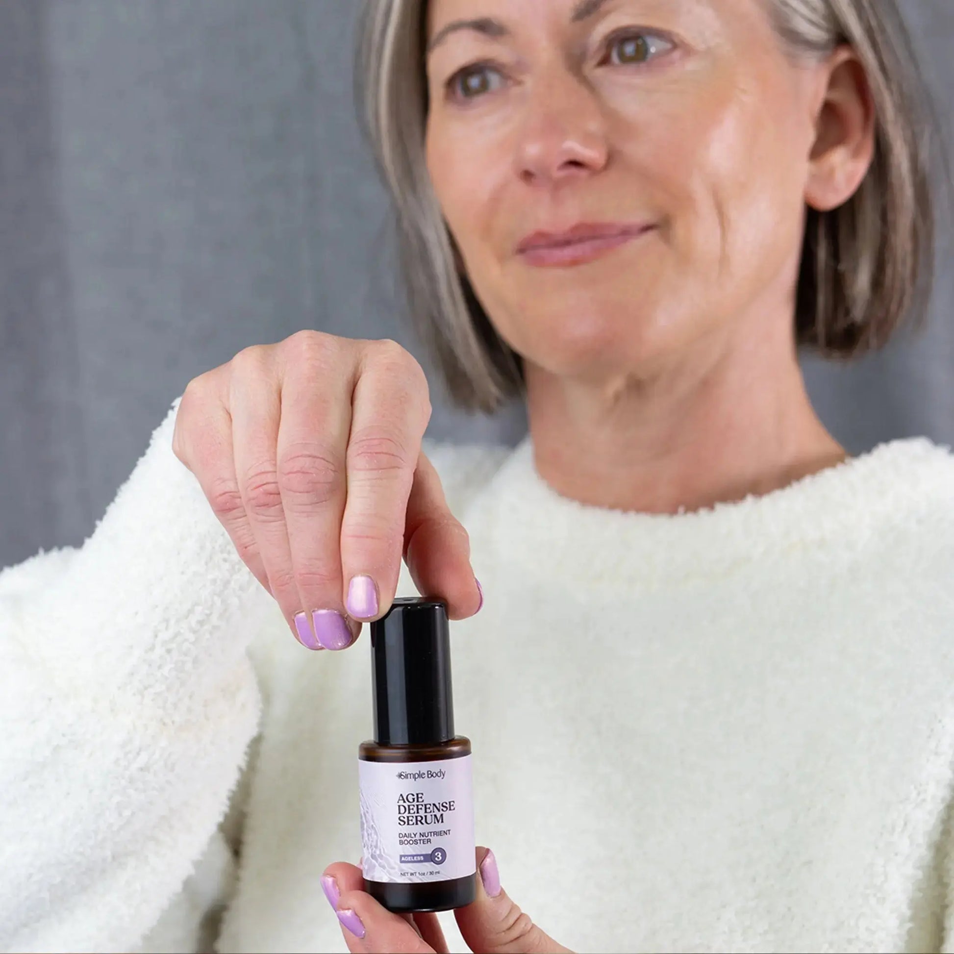 Woman holding a jar of Simple Body Age Defense Serum with a gray background