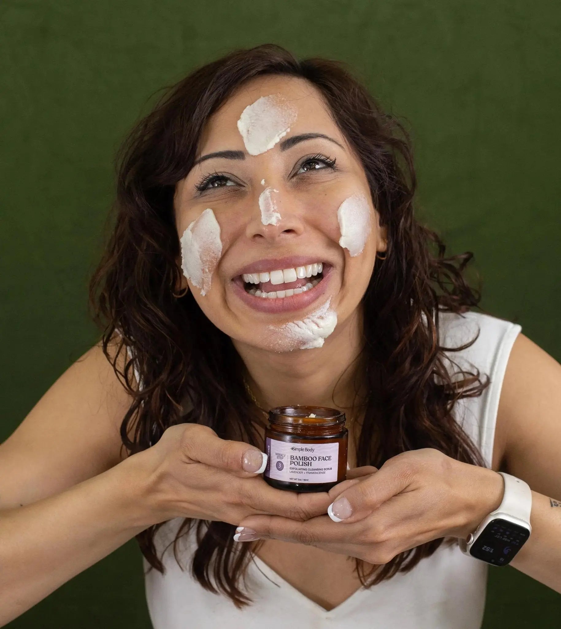 Woman applying Lavender Bamboo Face Polish to her face while holding a jar of Simple Body Bamboo Face Polish looking up and smiling.