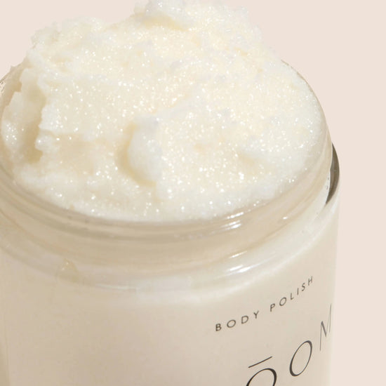 An open jar of Bloom Bath Co. body polish showing a creamy white, textured product inside, set against a solid pale peach background.