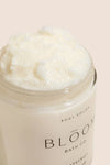 An open jar of Bloom Bath Co. body polish showing a creamy white, textured product inside, set against a solid pale peach background.