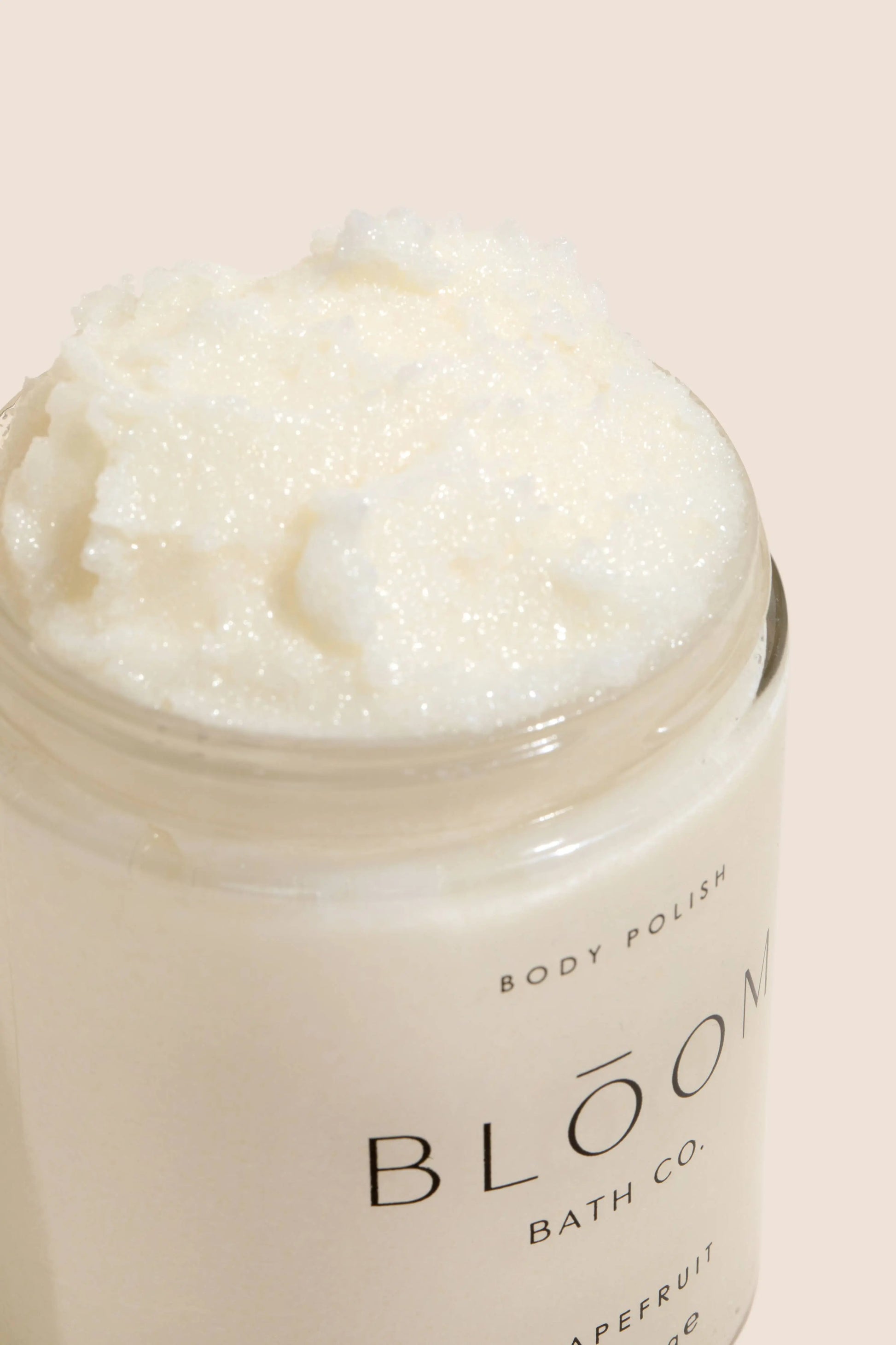 An open jar of Bloom Bath Co. body polish showing a creamy white, textured product inside, set against a solid pale peach background.