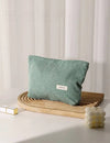 Green corduroy cosmetic bag on a wooden tray with a white curtain background