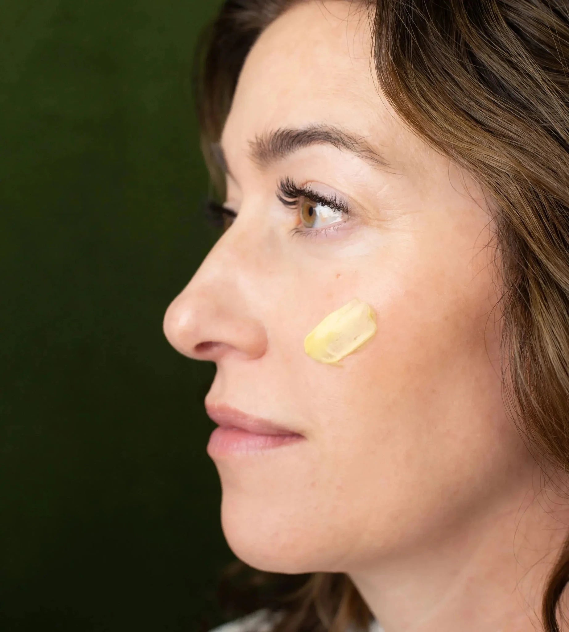A woman showing a swatch of Simple Body Beauty Balm on her cheek with a green background