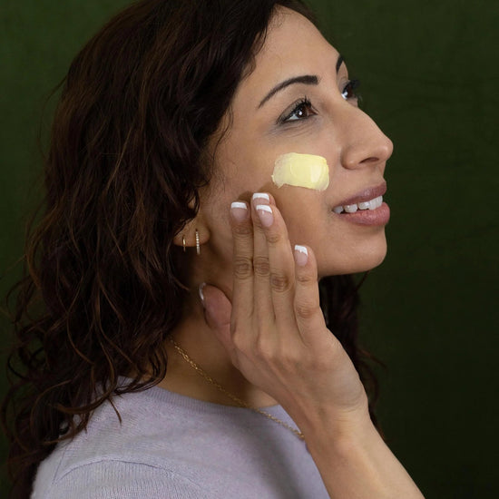 A woman looking sideways applying Simple Body Beauty Balm to her cheek which is a yellow, creamy texture.