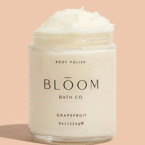 A clear plastic jar of Bloom Bath Co. Grapefruit body polish with white cream visible at the top, set against a solid peach background.