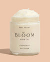 A clear plastic jar of Bloom Bath Co. Grapefruit body polish with white cream visible at the top, set against a solid peach background.