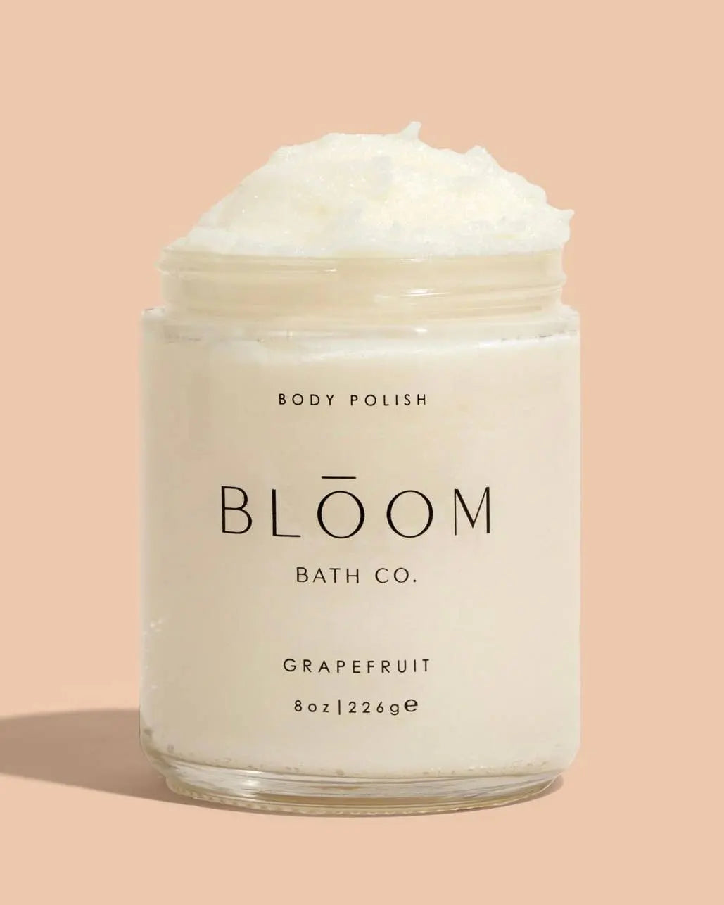 A clear plastic jar of Bloom Bath Co. Grapefruit body polish with white cream visible at the top, set against a solid peach background.