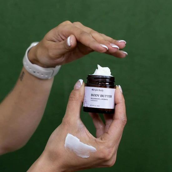 A woman getting ready to scoop some Simple Body, Body Butter in lavender out of a 1oz jar with a purple label and showing an off-white creamy thick texture product swipe on her hand.