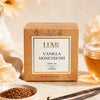 A box of LUMI Vanilla Honeybush herbal tea sits on a light surface with a glass of amber tea, vanilla beans, and loose tea seeds nearby. The box is tan with a white label and gold text.