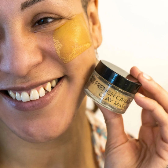 A young women models a Simple Body Clay Face Mask as she holds it against her cheek, smiling with a white background.