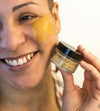 A young women models a Simple Body Clay Face Mask as she holds it against her cheek, smiling with a white background.