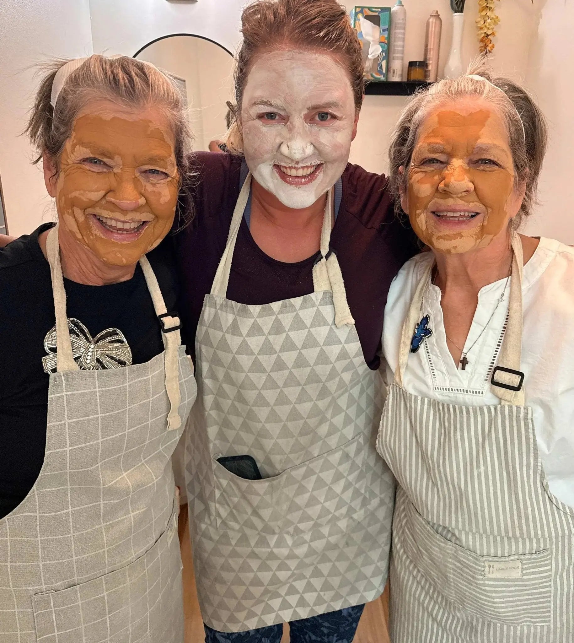 Three people with Simple Body Clay face masks on, wearing aprons, in a bathrom setting.