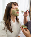 A woman applies a Simple Body French Green Clay Mask to another woman with long dark hair in a white robe.