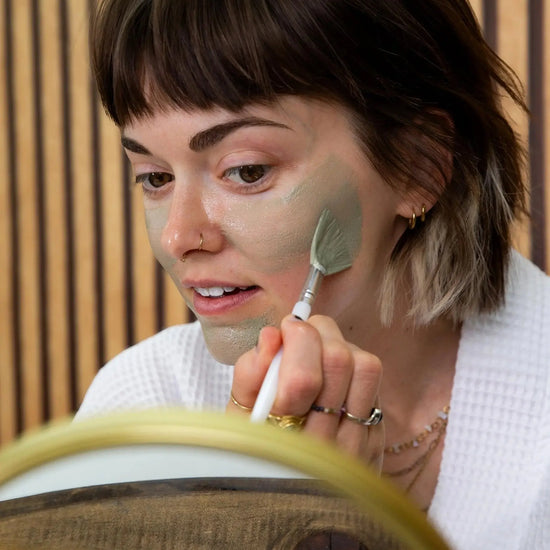 A woman applying a Simple Body French Green Clay Mask looking at a gold rimmed mirror with a wood wall backdrop.