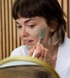 A woman applying a Simple Body French Green Clay Mask looking at a gold rimmed mirror with a wood wall backdrop.