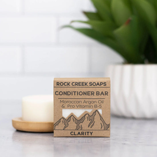 Rock Creek Soaps conditioner bar packaging on a surface with a candle and plant in the background