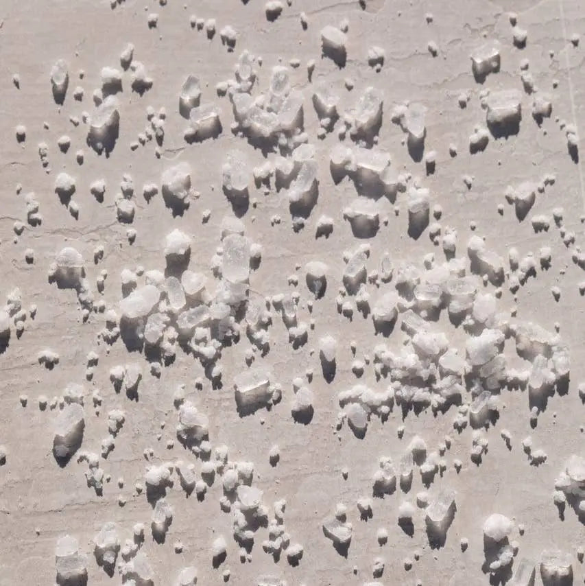 A close-up texture shot of coarse white bath salt crystals scattered across a light beige surface under bright sunlight.