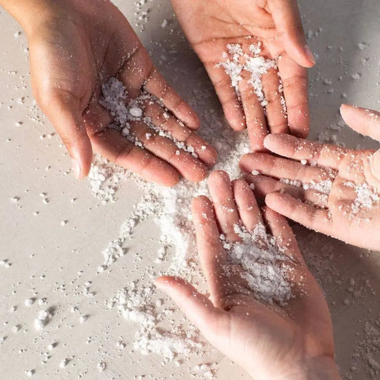 Four hands holding small amounts of white bath soak on a light surface