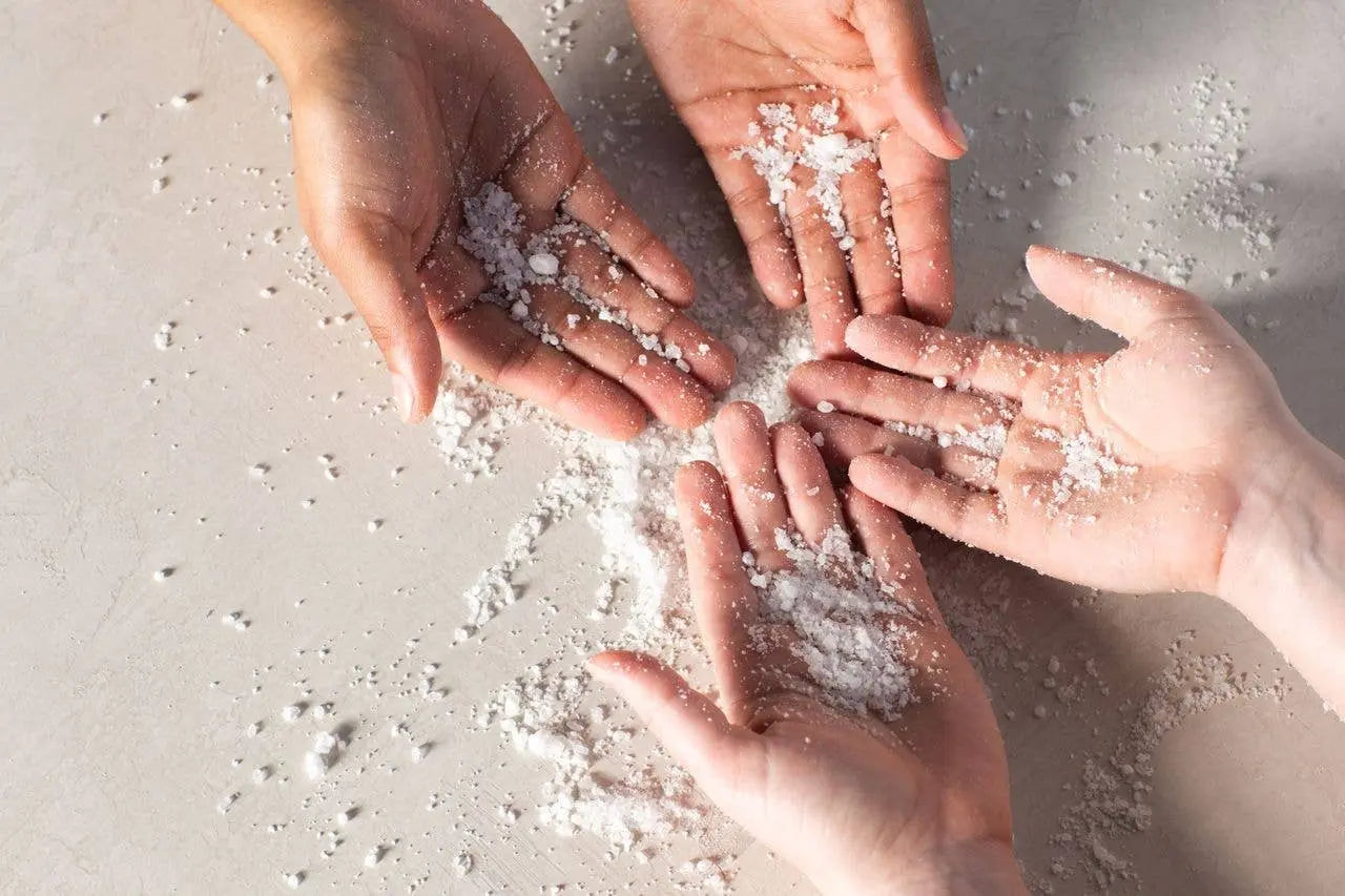 Four hands holding small amounts of white bath soak on a light surface
