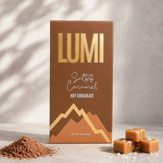 A box of Lumi Salted Caramel Hot Chocolate candy stands upright on a light stone surface with soft shadow overlays. The packaging is brown with metallic gold lettering and mountain graphics.
