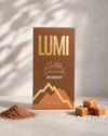 A box of Lumi Salted Caramel Hot Chocolate candy stands upright on a light stone surface with soft shadow overlays. The packaging is brown with metallic gold lettering and mountain graphics.