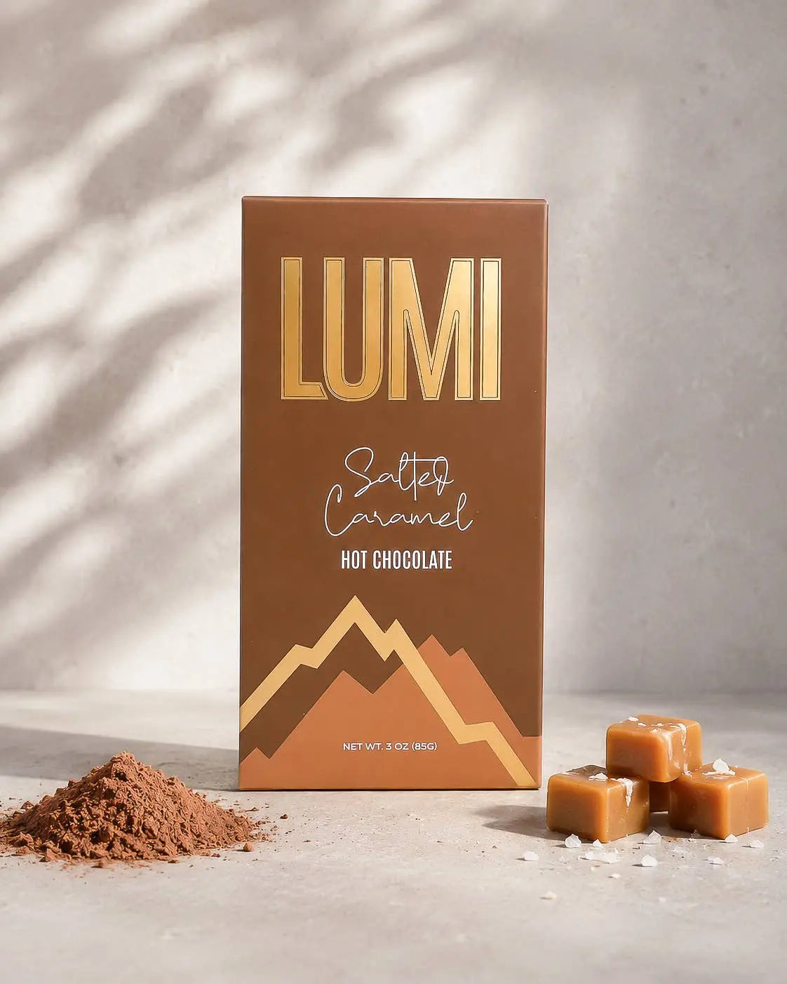 A box of Lumi Salted Caramel Hot Chocolate candy stands upright on a light stone surface with soft shadow overlays. The packaging is brown with metallic gold lettering and mountain graphics.