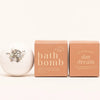 A white bath bomb by Ginger June in Daydream with herbs on top is shown next to two peach-colored boxes, one labeled 'bath bomb' and the other 'day dream', on a clean white background.