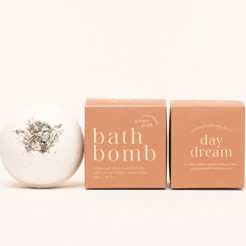 A white bath bomb by Ginger June in Daydream with herbs on top is shown next to two peach-colored boxes, one labeled 'bath bomb' and the other 'day dream', on a clean white background.